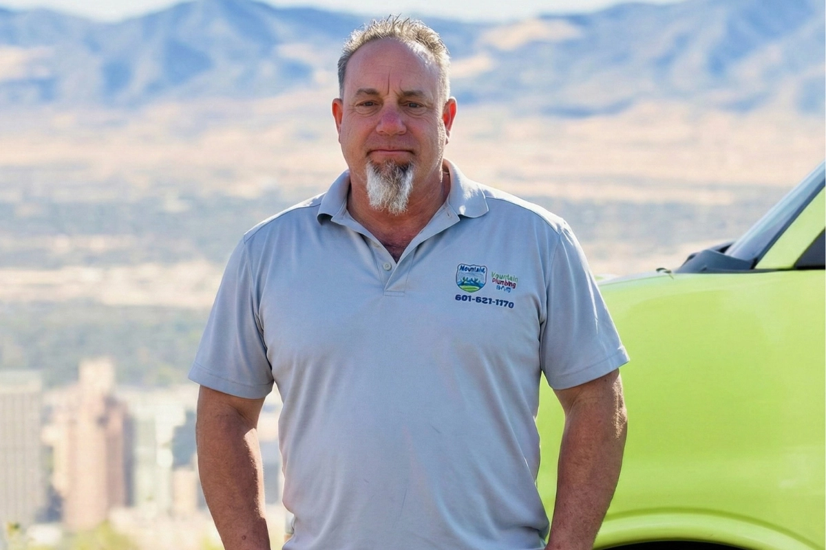 John stands confidently in front of a green vehicle, wearing a light gray polo shirt and khaki shorts. Mountains and a city skyline are visible behind him.